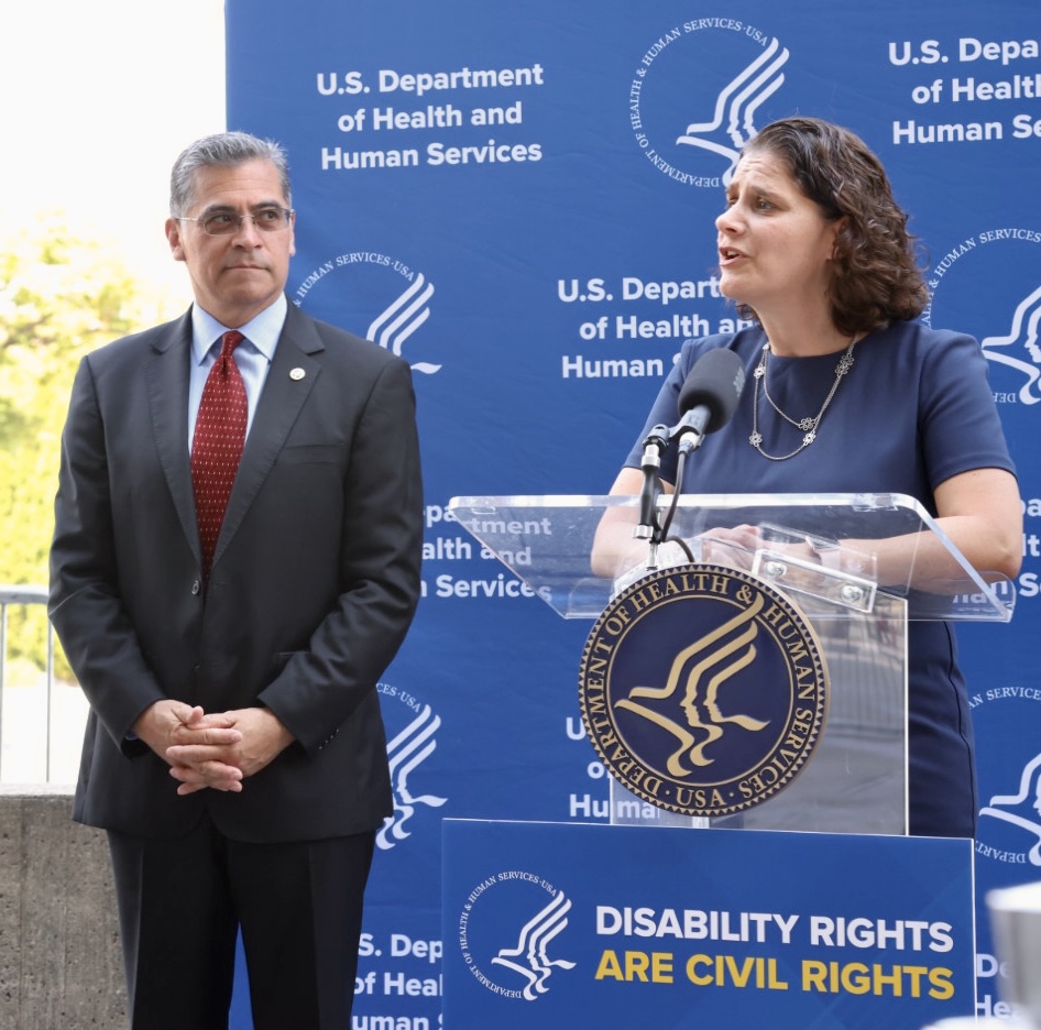 Secretary Xavier Becerra, who has short gray hair and is wearing a black suit with a blue shirt and red tie and glasses, stands next to Alison Barkoff, who has medium-length curly brown hair and is wearing a blue dress and is speaking from a clear lectern. They are standing in front of a blue background on which the words "U.S. Department of Health and Human Services" and the HHS seal are printed in a repeating pattern.