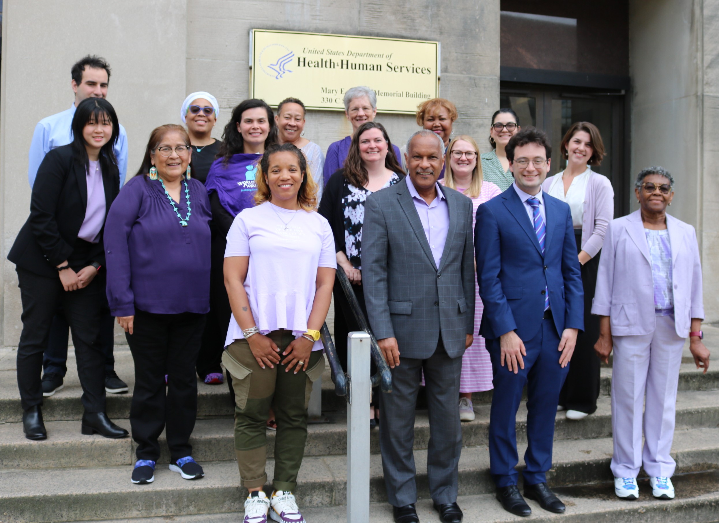 16 people wearing various shades of purple in front of a HHS building